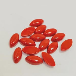 May include: A collection of small, smooth, marquise-shaped beads in a vibrant, solid red color. Each bead features a polished, glossy finish with a pointed oval silhouette, arranged in a loose cluster on a plain, neutral-toned surface.