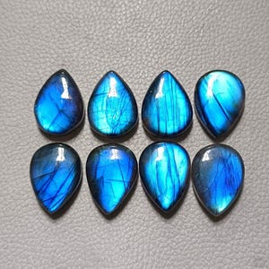 May include: Eight teardrop-shaped labradorite gemstones arranged in two rows of four. Each stone features a polished surface with a vibrant blue iridescent flash and visible internal fractures, displaying a smooth, rounded base and a pointed top.