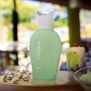 May include: A light green perfume bottle with a white cap, labeled "EXTREMELY UNIQUE PISTA." The bottle is on a table with pistachios and a dessert in the background. The bottle has a unique shape.