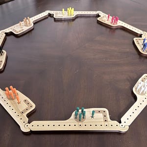 K&ouml;nnte beinhalten: Ein achteckiges Cribbage-Brett aus Holz mit Scharnierabschnitten. Das Brett enth&auml;lt verschiedene S&auml;tze farbiger Stifte, darunter gelb, rosa, blau, wei&szlig;, gr&uuml;n, orange und schwarz. Das Brett liegt auf einer dunkelbraunen Oberfl&auml;che.
