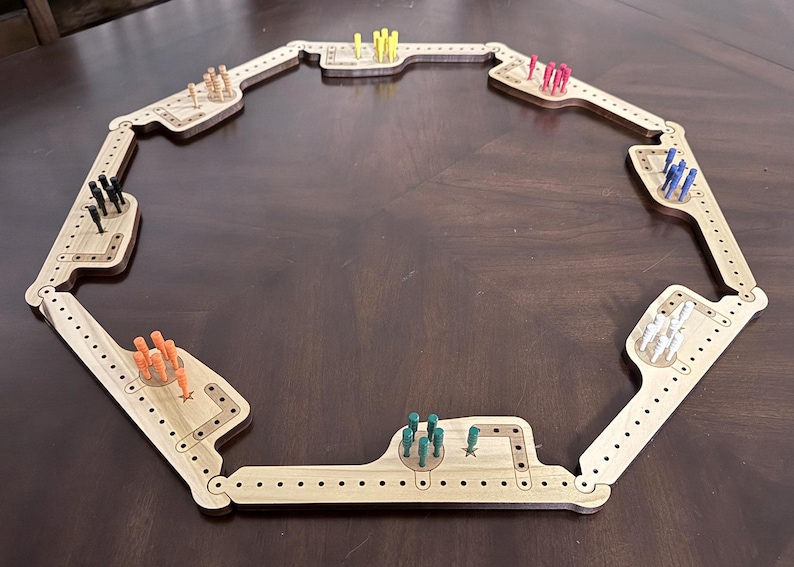K&ouml;nnte beinhalten: Ein achteckiges Cribbage-Brett aus Holz mit Scharnierabschnitten. Das Brett enth&auml;lt verschiedene S&auml;tze farbiger Stifte, darunter gelb, rosa, blau, wei&szlig;, gr&uuml;n, orange und schwarz. Das Brett liegt auf einer dunkelbraunen Oberfl&auml;che.