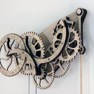 May include: Wooden wall clock featuring a gear mechanism design. The clock has a light wood finish with black accents, showcasing multiple interlocking gears and a pendulum. The clock's face displays numbers, and the overall design is intricate and decorative.