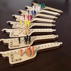 May include: Wooden cribbage boards with various colored pegs. The boards are light brown with a unique shape and a series of holes for peg placement. Pegs are in colors like red, blue, yellow, green, black, and orange.