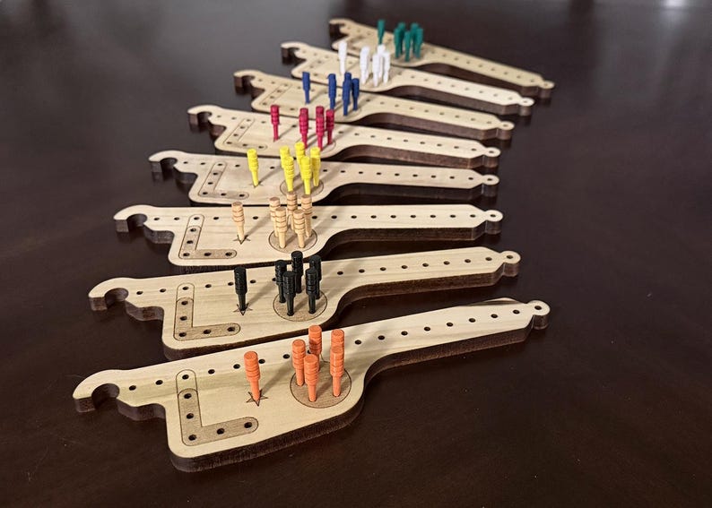 K&ouml;nnte beinhalten: Holz-Cribbage-Boards mit verschiedenfarbigen Stiften. Die Boards sind hellbraun mit einer einzigartigen Form und einer Reihe von L&ouml;chern f&uuml;r die Stiftplatzierung. Stifte sind in Farben wie Rot, Blau, Gelb, Gr&uuml;n, Schwarz und Orange.