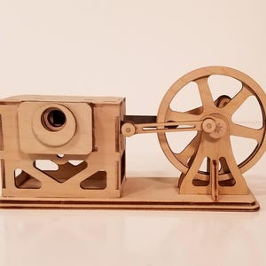 May include: A wooden model of a mechanical device, featuring a rectangular housing with a circular opening and a large spoked wheel connected by a rod. The model is constructed from light-colored wood.