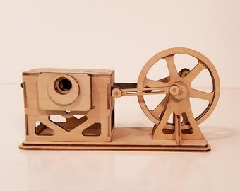 May include: A wooden model of a mechanical device, featuring a rectangular housing with a circular opening and a large spoked wheel connected by a rod. The model is constructed from light-colored wood.