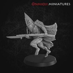 以下が含まれることがあります： ライフルに似た武器を持った、鎧を着た戦士のグレースケールミニチュアフィギュア。フィギュアは尖ったヘルメットと円形の切り抜きのあるケープを持っています。ベースは丸く、上部には「ONMIOJI MINIATURES」という文字があります。