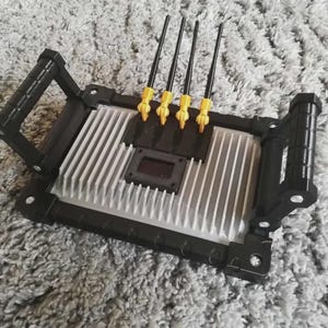 Può includere: Un dispositivo elettronico nero con una superficie argentata e scanalata e quattro antenne nere con punte gialle. Il dispositivo ha una forma rettangolare con maniglie nere su ogni lato. Il dispositivo è su una superficie grigia e strutturata.