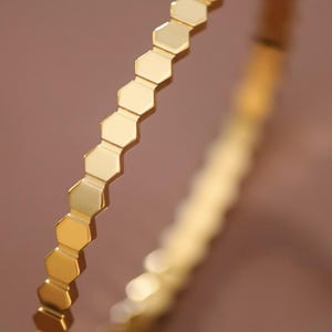 May include: A gold-colored bangle bracelet with a honeycomb design. The bracelet is made of a shiny, reflective metal and has a series of hexagonal shapes that run around the entire circumference. The bracelet is shown against a blurred brown background.