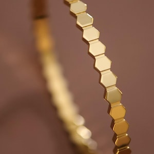 May include: A gold-colored bracelet with a honeycomb design. The bracelet is made of a shiny, reflective metal and features a series of hexagonal shapes. The bracelet is shown against a blurred brown background.
