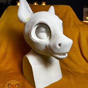 May include: A white, textured, foam-based animal mask base featuring large, rounded ears, a snout with a defined nose, and wide eye sockets. The mask is mounted on a smooth, white, neck-and-shoulder display stand, showcasing a blank, unpainted surface.