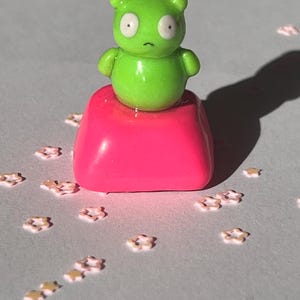 May include: A small, green cartoon character figurine with large white eyes and a downturned mouth sits atop a bright pink, rectangular base. Scattered around are small, star-shaped decorations with gold accents.