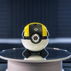 May include: A white, black, and yellow spherical device with a silver center, resting on a reflective surface. The top section is yellow and black, while the bottom is white. The device has a circular silver element in the center.