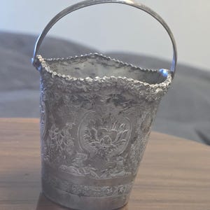 May include: A small, silver-colored bucket with a curved handle. The bucket features intricate floral and foliate designs around the body. The rim has a textured, scalloped edge. The bucket is likely made of metal.