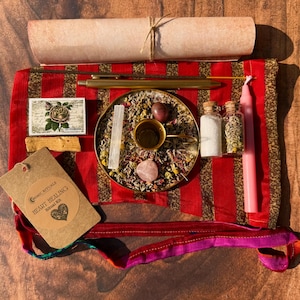 Könnte beinhalten: Ein Herzheilungs-Kit mit einem rot-gold gestreiften Tuch, einer Schriftrolle, Räucherstäbchen, einem Messingtablett mit Kräutern und Kristallen, zwei kleinen Glasflaschen, einer rosa Kerze und einem Etikett mit "Heart Healing".