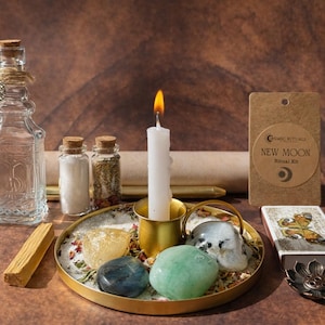 Pode incluir: Um kit ritual contendo uma garrafa de vidro transparente com um pingente de lua, dois frascos pequenos com pó branco e ervas, uma vela branca acesa num suporte dourado, uma bandeja dourada com três pedras polidas e uma etiqueta NEW MOON.