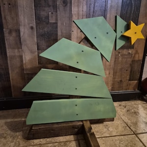 Könnte beinhalten: Ein grüner Weihnachtsbaum aus Holz, der aus abgewinkelten Brettern besteht. Er hat einen gelben Stern an der Spitze und einen dunkelbraunen Holzfuß und -stamm. Der Baum steht auf einem gefliesten Boden. Die Höhe beträgt ca. 100 cm.