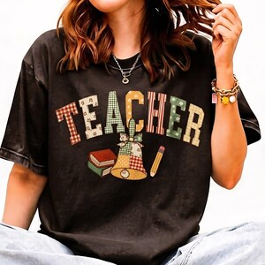 Może przedstawiać: Czarny t-shirt z krótkim rękawem z napisem TEACHER wykonanym dużymi, wielokolorowymi literami blokowymi z wzorami. Poniżej tekstu znajduje się grafika przedstawiająca dzwonek szkolny, stos dwóch książek i żółty ołówek w stylu patchworkowym.