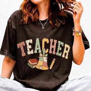 Puede incluir: Una camiseta de manga corta en color gris carbón oscuro con la palabra TEACHER en letras mayúsculas grandes, estampadas y multicolores. Debajo del texto hay una ilustración gráfica de dos libros apilados, una campana y un lápiz amarillo.