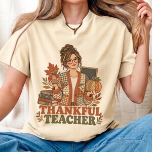 以下が含まれることがあります： クリーム色のTシャツで、メガネをかけてお団子ヘアの人物がパッチワーク柄のカーディガンを着ているグラフィックが描かれています。人物は本と黒板の横で2つの柄付きカボチャを持っています。下にTHANKFUL TEACHERという文字があります。