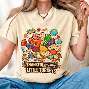 Op de afbeelding: Een crèmekleurig t-shirt met een centrale afbeelding van een kalkoen met kleurrijke, gedessineerde veren. Rondom de kalkoen staan schoolthema-iconen zoals een bus, boeken en globes. De tekst luidt: "THANKFUL for my LITTLE TURKEYS."
