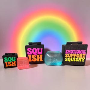 May include: Two black square boxes with hinges, one labeled SQUISH in pink and orange, the other EMOTIONAL SUPPORT SQUISH in pink and orange. Beside them are two translucent squishy cubes, one pink and one blue, each embossed with the text NEED DOH.