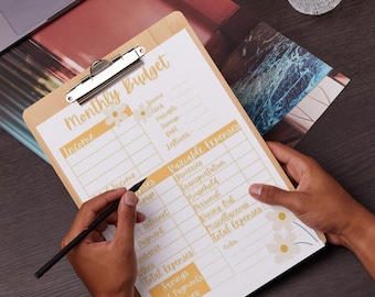 Puede incluir: Un portapapeles de madera con un formulario impreso de "Presupuesto Mensual". El formulario incluye secciones para ingresos y gastos variables, con detalles florales. Una persona sostiene un bolígrafo negro, lista para escribir en el formulario. Un portátil y una planta están en el fondo.