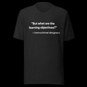 Könnte beinhalten: Dunkelgraues T-Shirt mit weißem Text: „Aber was sind die Lernziele?“ und „~ Instructional Designer“. Das Shirt hat einen Rundhalsausschnitt und kurze Ärmel.