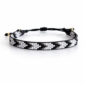 May include: Woven bracelet with a black, white, and gray geometric pattern. The bracelet has a black cord with adjustable knots and gold-colored accents. A stylish accessory for everyday wear.