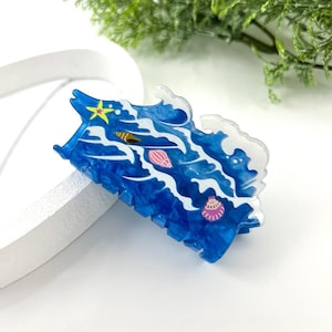 Könnte beinhalten: Eine blau-weiße Haarspange mit Wellenmuster. Die Spange zeigt einen gelben Seestern und mehrere Muscheln in Rosa, Gold und Weiß. Die Spange ist etwa 7,6 cm lang.