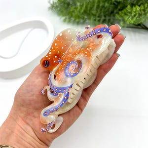 Puede incluir: Una pinza para el pelo decorativa con forma de pulpo. La pinza es naranja y blanca con detalles azules en los tent&aacute;culos. La pinza est&aacute; hecha de un material brillante y est&aacute; dise&ntilde;ada para sujetar el pelo en su sitio. Un accesorio para el pelo &uacute;nico.