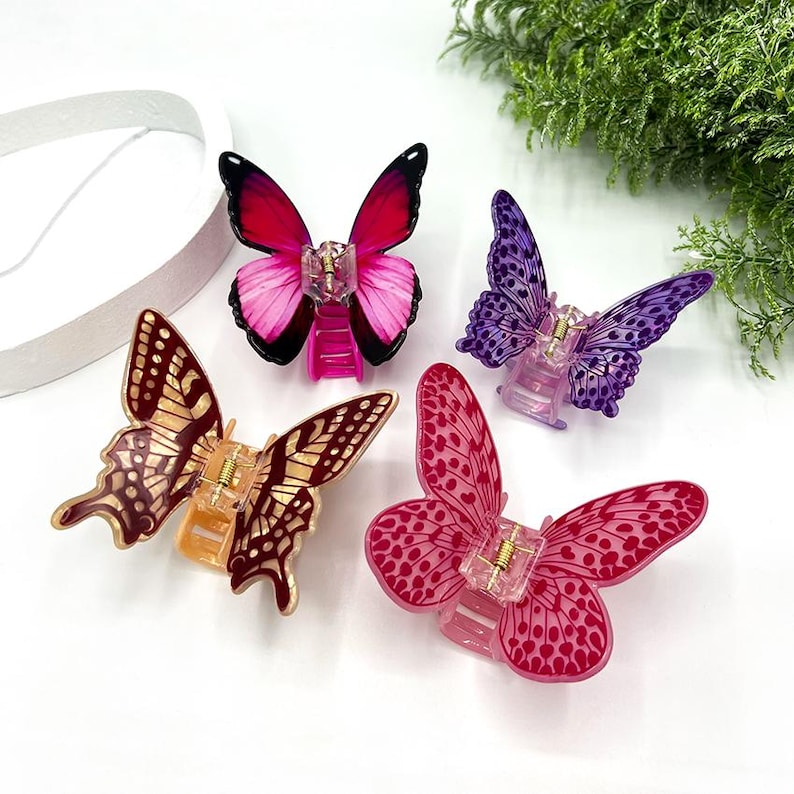 May include: Four butterfly hair clips in shades of pink, purple, and gold. Each clip features a detailed butterfly wing design and a clear plastic clasp. The clips are arranged on a white surface with greenery in the background.