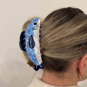 Pode incluir: Uma presilha de cabelo em forma de peixe azul e branco. A presilha apresenta um design de peixe sorridente com cauda e barbatanas azuis escuras. O acess&oacute;rio de cabelo &eacute; feito de um material brilhante e &eacute; preso em um coque.