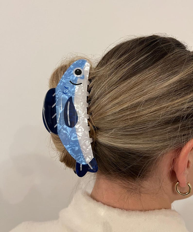 Pode incluir: Uma presilha de cabelo em forma de peixe azul e branco. A presilha apresenta um design de peixe sorridente com cauda e barbatanas azuis escuras. O acess&oacute;rio de cabelo &eacute; feito de um material brilhante e &eacute; preso em um coque.