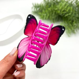 May include: A vibrant pink butterfly hair claw clip with black accents. The clip features a detailed butterfly wing design in shades of pink and black, with a bright pink claw mechanism.