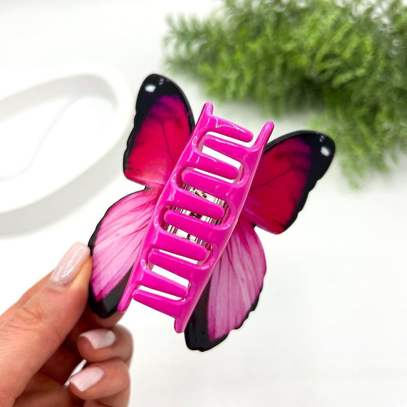 May include: A vibrant pink butterfly hair claw clip with black accents. The clip features a detailed butterfly wing design in shades of pink and black, with a bright pink claw mechanism.