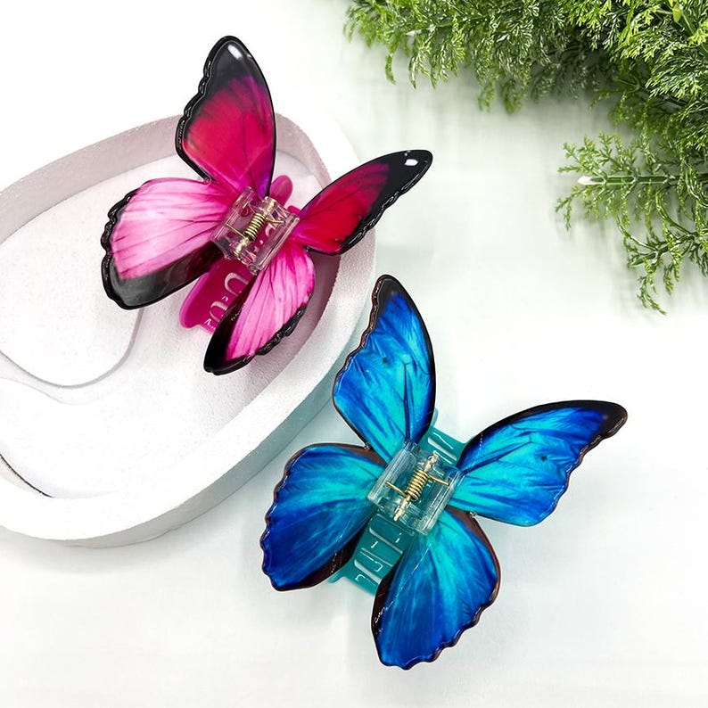 May include: Two butterfly-shaped hair clips. One clip features pink and red wings with black edges, the other has blue wings with black edges. Both clips have a clear plastic base with a gold-colored spring.