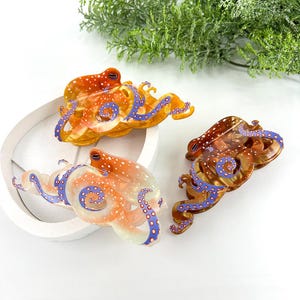 Puede incluir: Tres pinzas para el pelo con forma de pulpo en tonos naranja, marr&oacute;n y transparente. Las pinzas presentan lunares blancos y tent&aacute;culos azules. Las pinzas est&aacute;n dispuestas sobre una superficie blanca con follaje verde en el fondo.