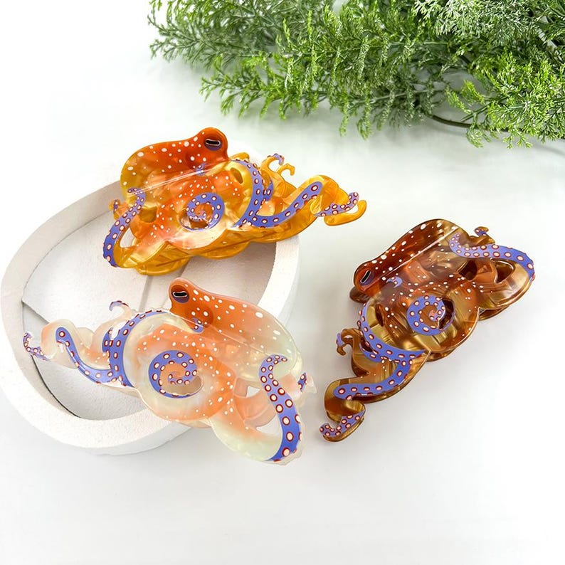 Puede incluir: Tres pinzas para el pelo con forma de pulpo en tonos naranja, marr&oacute;n y transparente. Las pinzas presentan lunares blancos y tent&aacute;culos azules. Las pinzas est&aacute;n dispuestas sobre una superficie blanca con follaje verde en el fondo.