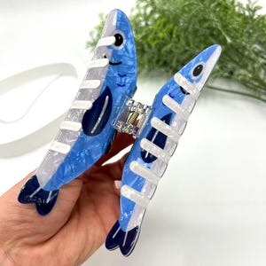 Pode incluir: Uma presilha de cabelo azul e branca em forma de dois peixes. A presilha tem um efeito marmorizado com detalhes em azul escuro e um fecho prateado. Os peixes t&ecirc;m olhos pretos e est&atilde;o virados em dire&ccedil;&otilde;es opostas.