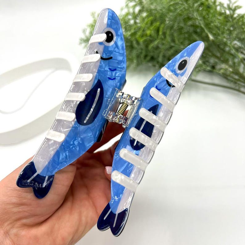 Pode incluir: Uma presilha de cabelo azul e branca em forma de dois peixes. A presilha tem um efeito marmorizado com detalhes em azul escuro e um fecho prateado. Os peixes t&ecirc;m olhos pretos e est&atilde;o virados em dire&ccedil;&otilde;es opostas.