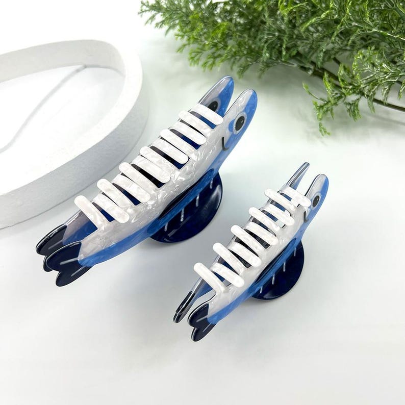 Pode incluir: Duas presilhas de cabelo em forma de peixe. As presilhas s&atilde;o azuis, brancas e pretas, com um design de peixe. As presilhas t&ecirc;m uma estrutura branca em forma de pente na parte superior. As presilhas est&atilde;o em uma superf&iacute;cie branca com vegeta&ccedil;&atilde;o ao fundo.