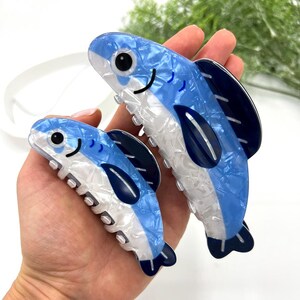 Pode incluir: Duas presilhas de cabelo em forma de peixe azul e branco. As presilhas t&ecirc;m um padr&atilde;o marmorizado, olhos pretos e uma boca sorridente. As barbatanas e a cauda s&atilde;o azul escuro. As presilhas s&atilde;o seguradas em uma m&atilde;o.