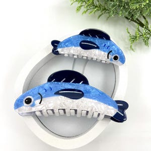 Pode incluir: Duas presilhas de cabelo em forma de peixe azul e branco. As presilhas t&ecirc;m uma barbatana e cauda azul escuro, com um corpo branco e azul. As presilhas est&atilde;o numa bandeja oval branca, com folhagem verde no fundo.