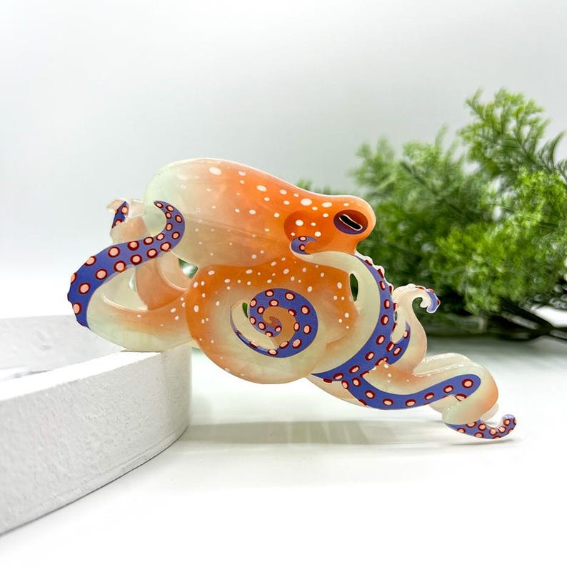 Puede incluir: Una colorida escultura de pulpo de cristal. El pulpo presenta un cuerpo naranja con puntos blancos, tent&aacute;culos azules y morados y una parte inferior de color crema. La escultura est&aacute; sobre un fondo blanco con vegetaci&oacute;n.