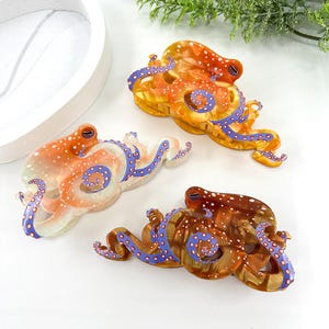 Puede incluir: Tres pinzas para el pelo con forma de pulpo en tonos naranja, marr&oacute;n y transparente. Cada pinza presenta un patr&oacute;n &uacute;nico de puntos blancos y tent&aacute;culos azules con detalles morados. Las pinzas est&aacute;n dise&ntilde;adas con un acabado brillante.