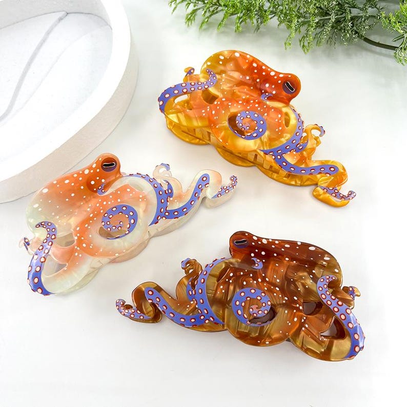 Puede incluir: Tres pinzas para el pelo con forma de pulpo en tonos naranja, marr&oacute;n y transparente. Cada pinza presenta un patr&oacute;n &uacute;nico de puntos blancos y tent&aacute;culos azules con detalles morados. Las pinzas est&aacute;n dise&ntilde;adas con un acabado brillante.