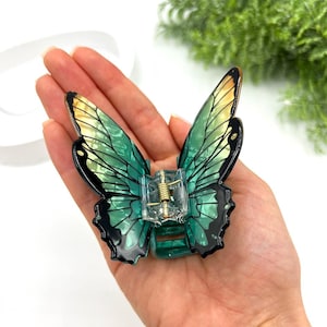 May include: A butterfly-shaped hair clip with translucent green and yellow wings, outlined in black. The clip has a clear, plastic base with a gold-colored metal spring mechanism. The wings are detailed with veining.