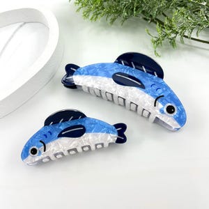 Pode incluir: Duas presilhas de cabelo em forma de peixe. As presilhas s&atilde;o azuis e brancas com detalhes em azul escuro, assemelhando-se a um peixe. As presilhas t&ecirc;m um acabamento texturizado e perolado e um fecho tipo garra. As presilhas est&atilde;o sobre uma superf&iacute;cie branca.