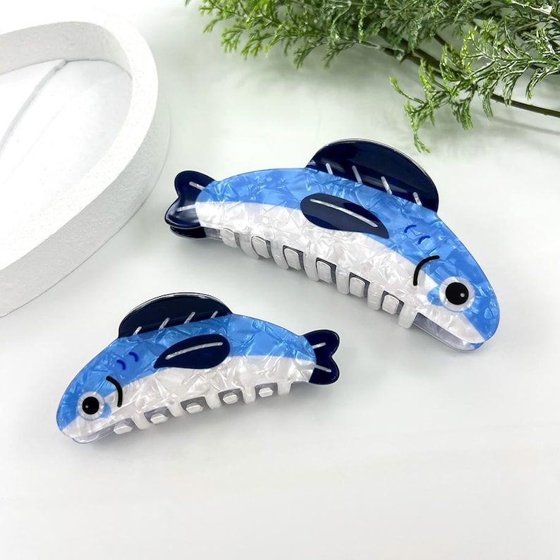 Pode incluir: Duas presilhas de cabelo em forma de peixe. As presilhas s&atilde;o azuis e brancas com detalhes em azul escuro, assemelhando-se a um peixe. As presilhas t&ecirc;m um acabamento texturizado e perolado e um fecho tipo garra. As presilhas est&atilde;o sobre uma superf&iacute;cie branca.